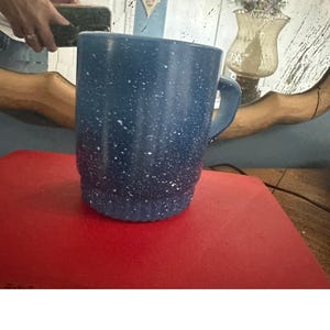 Puede incluir: Una taza azul moteada con asa. La taza tiene una base azul oscuro que se desvanece a un azul más claro hacia la parte superior. Las motas blancas están dispersas por la superficie. La taza está sobre una superficie roja.