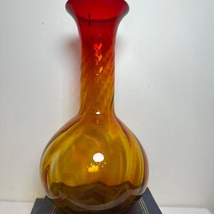 Może przedstawiać: Ozdobny szklany wazon z pękatą podstawą i rozkloszowaną, czerwoną krawędzią. Wazon przechodzi od głębokiego bursztynu u podstawy do żywego pomarańczu i czerwieni u góry. Szkło ma wzór wirowy.