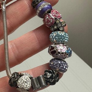 Puede incluir: Una pulsera de abalorios plateada con varias cuentas decorativas. Las cuentas incluyen esferas moradas, rosas y azules con incrustaciones de cristal, dijes en forma de corazón y cuentas en forma de rosa negra. La pulsera se sostiene en una mano.