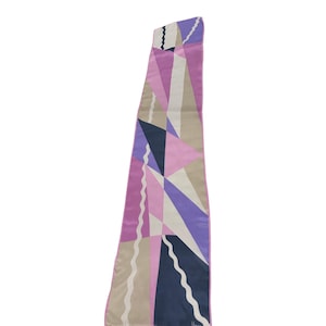 May include: A long, narrow scarf featuring a geometric pattern of triangles and shapes in shades of lavender, light pink, dark navy blue, and beige. The design includes thin, white, wavy vertical lines running across the fabric surface.