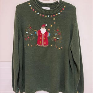 Può includere: Un maglione a maniche lunghe verde scuro con una figura ricamata di Babbo Natale in cappotto rosso, affiancato da due pini ricamati. Lo scollo è decorato con piccoli bottoni colorati e dettagli a forma di stella, con ulteriori stelle sparse attorno al motivo.