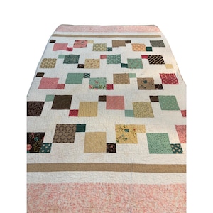 Puede incluir: Una colcha rectangular con fondo blanco y un patrón disperso de cuadrados de tela multicolor en tonos rosa, verde azulado, marrón, amarillo y estampados florales. La colcha está bordeada por bandas de color canela y tela floral rosa.