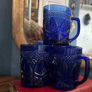 May include: Four cobalt blue glass mugs with a diamond and leaf pattern are stacked on a red book. Each mug has a handle. The mugs are reflecting in a gold-framed mirror.