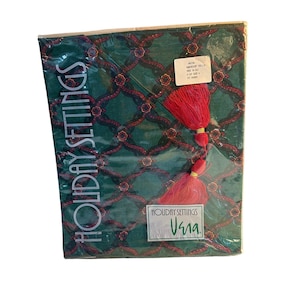 May include: A package of Holiday Settings placemats. The placemats feature a green background with a repeating pattern of red and gold floral designs. A red tassel is attached. The text "Holiday Settings" and "Vana" are visible.
