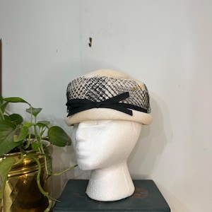 Puede incluir: Un sombrero vintage color crema con una superposición de red negra y un detalle de cinta negra. El sombrero se exhibe en una cabeza de maniquí blanca, colocada sobre una pila de libros verde oscuro. Se ve una maceta dorada con una planta verde.