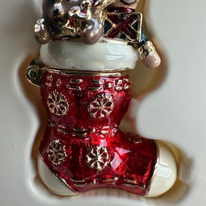 Puede incluir: Adorno de calcetín navideño con acabado rojo brillante y detalles de copos de nieve dorados. El calcetín está rematado con un puño blanco y presenta un soldadito de juguete y un osito de peluche. El adorno está sobre una base crema.