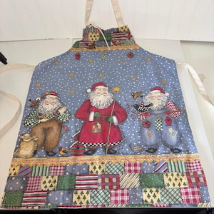 May include: A blue apron featuring a white polka dot pattern and three illustrated Santa Claus figures. The bottom border displays a colorful patchwork design of squares with various patterns. The apron includes cream-colored fabric neck and waist ties.