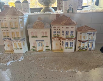 May include: A set of four ceramic house-shaped canisters with lids. Each canister features painted architectural details including windows, doors, and roofs in shades of blue, brown, and green on a cream-colored base. The structures vary in height.