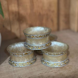 May include: A set of four decorative napkin rings. The rings are a pale gold color with a silver and gold filigree design around the top and bottom edges. The napkin rings are stacked on a wooden surface.