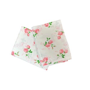 May include: Two folded white fabric napkins with a repeating pattern of pink roses and green leaves. The floral design is scattered across the fabric, creating a delicate and feminine aesthetic. The napkins are likely made of a soft material.