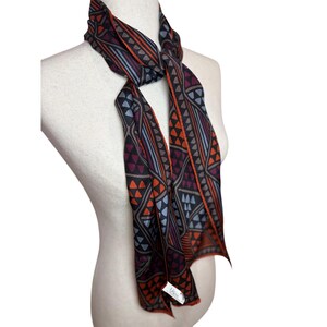 May include: A long, narrow scarf featuring a geometric pattern of triangles and lines in shades of black, dark orange, purple, and light blue. The scarf is tied around the neck of a mannequin and includes a small white brand tag at the bottom edge.
