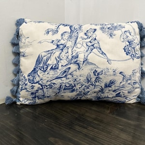 May include: A white throw pillow with blue and white toile print featuring a pastoral scene with figures. The pillow has blue tassel trim.