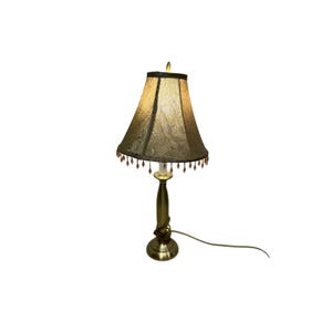 May include: A vintage-style table lamp with a gold-toned base and a patterned lampshade. The lampshade is a dark olive green color with a floral design and beaded fringe. The lamp has a cord and a lightbulb.