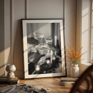 May include: Black and white framed print of a celebratory toast with crystal glasses. The image is displayed on a wooden floor, next to a white vase with orange flowers, and a small decorative vase.