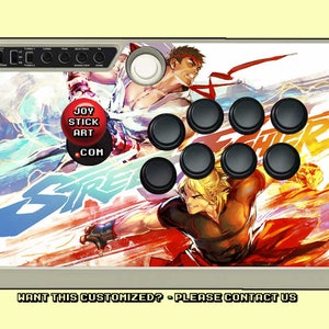 May include: A white and gray arcade joystick with a Street Fighter V design. The joystick features a white button with the text "JOY STICK ART .COM" and eight black buttons. The design features two characters from the Street Fighter video game series.