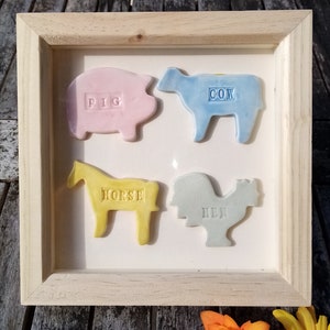 May include: A framed artwork featuring four ceramic farm animal shapes: a pink pig, a blue cow, a yellow horse, and a light blue hen. Each animal has its name embossed on its body. The artwork is set in a natural wood frame.