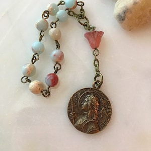 Op de afbeelding: Een blauwe en witte kralenrosary met een rode glazen bloemenhanger en een bronzen medaille met het profiel van een vrouw.