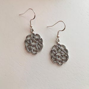May include: A pair of silver-colored Celtic knot earrings. Each earring features a circular, interwoven knot design suspended from a hook. The earrings are displayed on a white surface, with a pink flower and a string of pearls in the background.