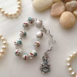 May include: White and blue floral bead bracelet with a silver charm of a woman in a long dress. The bracelet is on a white surface with white stones.