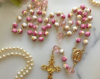 Puede incluir: Un rosario floral blanco y rosa con detalles dorados. El rosario tiene un crucifijo dorado y un colgante ovalado dorado con una representación de la Virgen María.