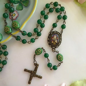 May include: Green jade bead rosary with a bronze Celtic cross and a bronze oval pendant featuring a religious image. The beads are accented with gold shamrocks.