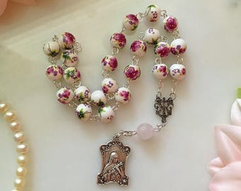 May include: A rosary featuring white ceramic beads adorned with purple floral designs. It includes a silver-tone crucifix and a pendant with a religious figure. A pink gemstone and a pearl necklace are also present.