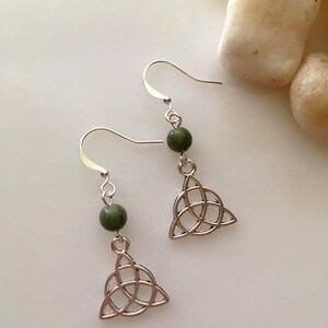 May include: A pair of silver-toned earrings featuring a Celtic knot design. Each earring has a green bead and a dangling, openwork triangular knot. The earrings are displayed on a white surface, with a few light-colored stones in the background.