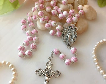 May include: A rosary with white beads adorned with pink floral patterns. The rosary features a silver crucifix and a silver centerpiece with a religious figure. A pearl bracelet is visible in the background.