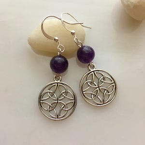 May include: A pair of silver-toned earrings featuring a Celtic knot design. Each earring has a round, deep purple bead and a circular pendant with an intricate knot pattern. The earrings hang from silver-colored ear wires.