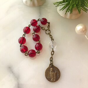 May include: A red glass beaded rosary with a clear glass bead and a bronze pendant featuring a religious image. The pendant has the text "SANCTUS LUCIA ORA PRO NOBIS".