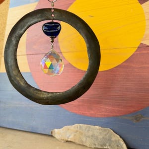 May include: A rustic metal ring wind chime with a faceted crystal ball and a dark blue glass bead. The wind chime hangs in front of a colorful wooden background with a yellow circle and red and blue stripes.