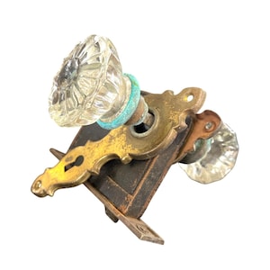May include: Antique door hardware set featuring two clear glass doorknobs with a ribbed design. The set includes a brass-colored escutcheon plate and a dark metal mortise lock, showcasing a vintage aesthetic.