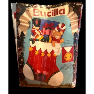 Könnte beinhalten: Ein Bucilla Jumbo-Filzstrumpf-Set in einer Kunststoffverpackung. Der rote Strumpf zeigt einen Spielzeugsoldaten, einen Teddybären und Geschenke. Der Text gibt 71 cm Diagonale und Bob an. Das Set enthält Filz, Pailletten, Perlen, Nadeln und Stickgarn.