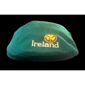 May include: A dark green flat cap featuring the word Ireland embroidered in white thread, accompanied by a gold-colored shamrock emblem positioned directly above the text. The cap has a rounded, structured crown and a short, curved brim.