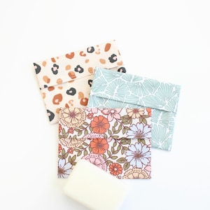 Op de afbeelding: Drie stoffen zeepzakjes met verschillende patronen: een tan luipaardprint, een lichtblauw bladontwerp en een bloemenpatroon met oranje, roze en blauwe bloemen. Een witte zeep ligt ervoor.