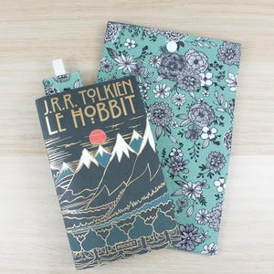 Peut inclure: Une couverture de livre et un marque-page assorti avec le titre "J.R.R. Tolkien Le Hobbit" et un motif de paysage de montagne. Une housse de livre à motifs floraux en teintes de sarcelle, blanc et noir est également présente. Le marque-page est attaché au livre.