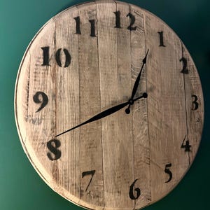 May include: A round wall clock constructed from vertical light-colored wooden planks. The clock face features large, black painted Arabic numerals from 1 to 12 and two thin black metal hands indicating the time. The wood grain is visible throughout.