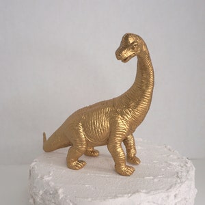 May include: A gold-painted dinosaur cake topper. The dinosaur is standing on a white frosted cake.