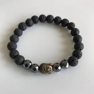 Puede incluir: Pulsera de cuentas de roca volcánica negra y hematita con un dije de cabeza de Buda dorado.