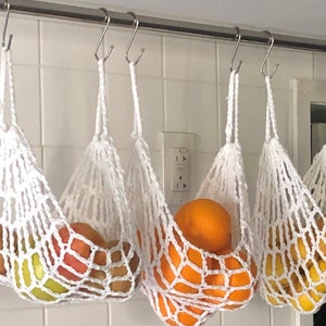 May include: Four white crocheted hanging produce bags filled with oranges and apples. The bags are hanging from hooks on a white tiled wall.