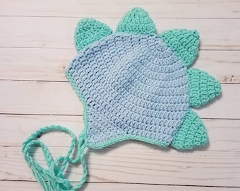May include: A handmade crochet dinosaur hat in light blue and teal. The hat features a rounded crown and teal spikes along the top. A teal tie with fringe hangs from the hat.