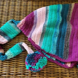 May include: A crocheted hooded scarf with a striped pattern in shades of pink, purple, teal, and gray. The scarf has a long tassel and a pom-pom at the end.