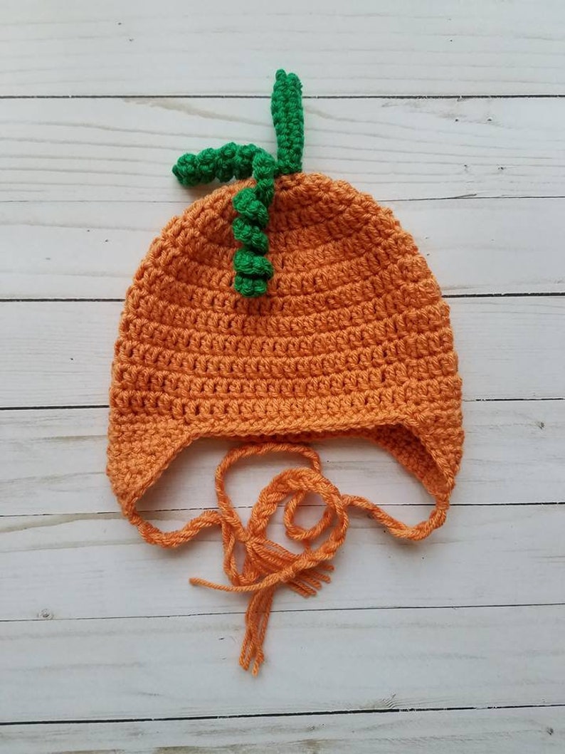 May include: A bright orange crocheted hat featuring a textured pumpkin-like design with a green stem and a curly green vine on top. The hat includes ear flaps and two long orange braided ties hanging from the bottom for securing the garment.