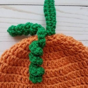 May include: A crocheted orange pumpkin hat featuring a textured surface with a green stem and a curly green vine attached to the top. The yarn work displays a consistent stitch pattern throughout the rounded crown of the headwear item.