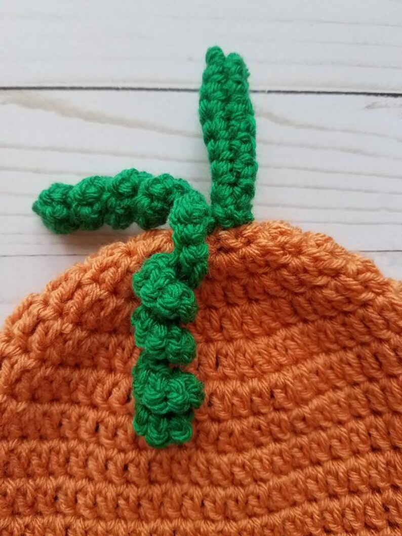 May include: A crocheted orange pumpkin hat featuring a textured surface with a green stem and a curly green vine attached to the top. The yarn work displays a consistent stitch pattern throughout the rounded crown of the headwear item.