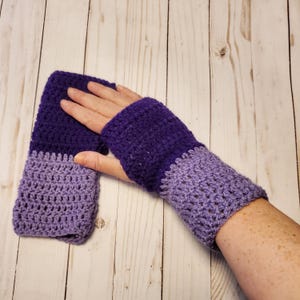 May include: Purple and lavender crocheted fingerless gloves. The gloves are made with a textured stitch and have a two-tone color scheme.