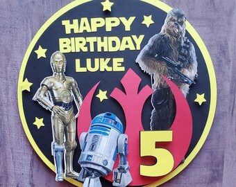 May include: A Star Wars-themed birthday cake topper featuring C-3PO, Chewbacca, and R2-D2. The black circle has yellow stars and the text "HAPPY BIRTHDAY LUKE". A red symbol and the number "5" are also included.