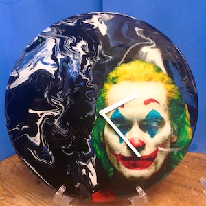 May include: A black and white marbled vinyl record with a Joker face printed on it. The Joker is wearing green hair and a red smile. The record is on a white stand.
