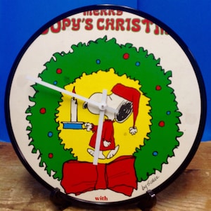 May include: A round, white clock with a black rim. The clock face features a cartoon image of Snoopy holding a candle in front of a green wreath with red and blue accents. The text "Merry Snoopy's Christmas with" is printed on the clock face. The clock has black hands and a black stand.