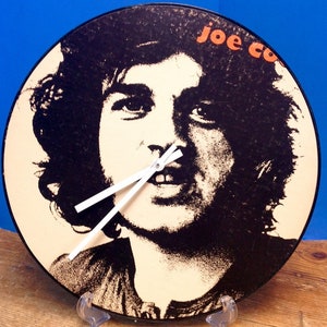 May include: A black vinyl record clock featuring a black and white image of Joe Cocker with the text "Joe Cocker" in orange and white. The clock has white hands.